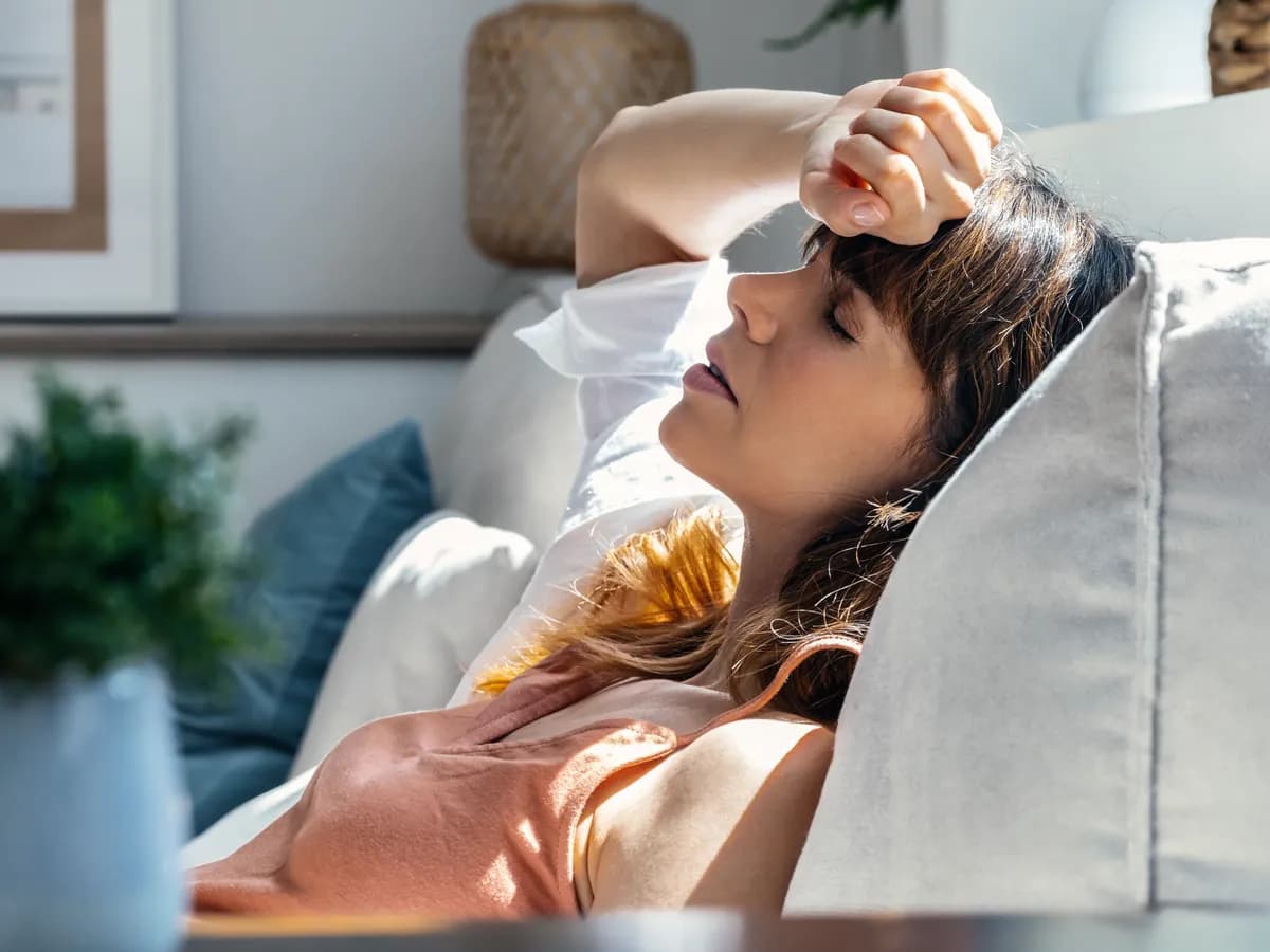 Woman lying back on her couch in afternoon light with her eyes closed and a hand on her forehead