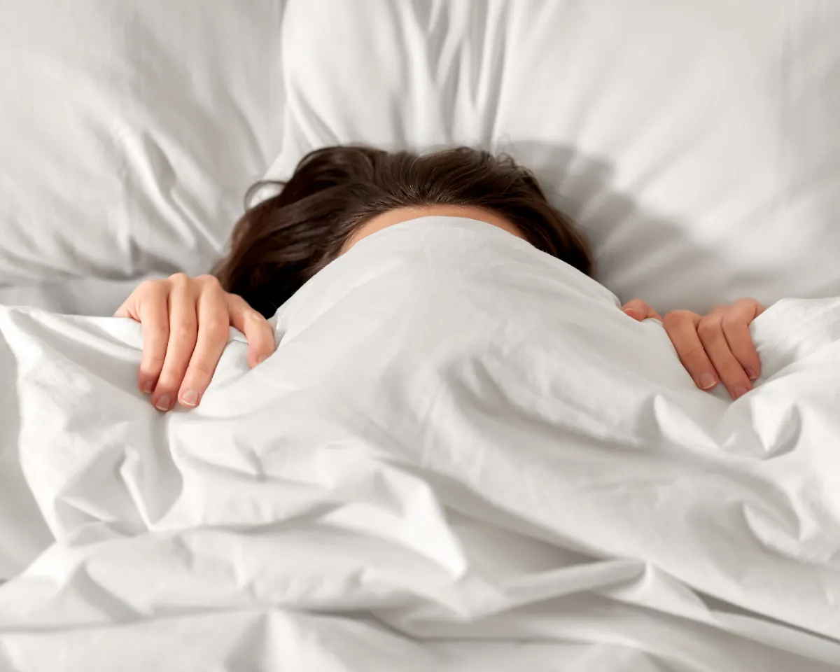 Woman lying in bed, pulling the duvet up over her face
