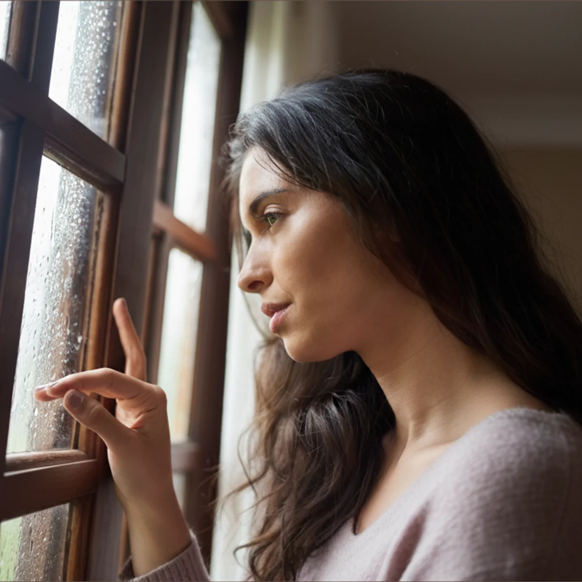 Woman gazing out a rainy window, sitting with quiet sadness on a grey day